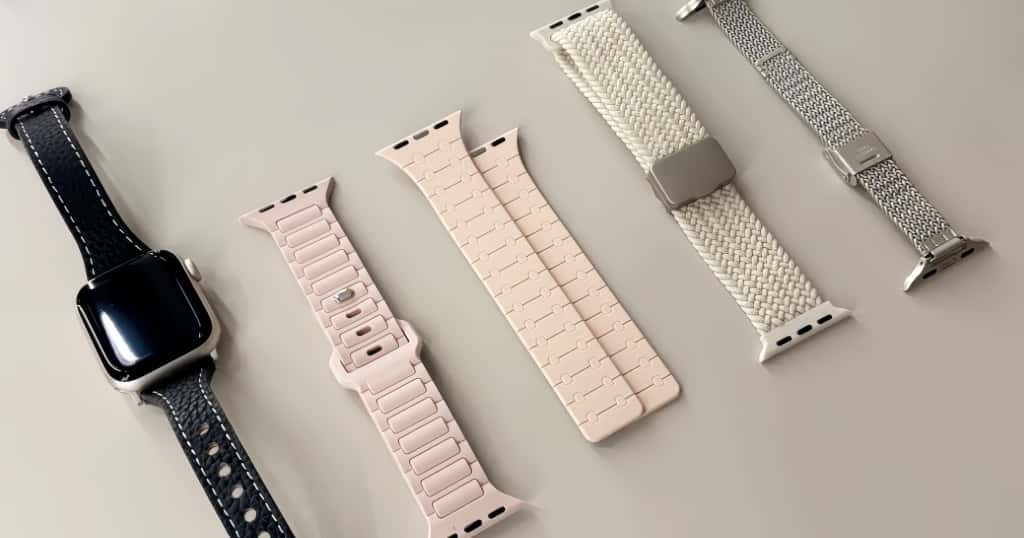Különböző anyagokból készült Apple Watch szíjak vannak elhelyezve egy asztalon, feltüntetve a szíjanyagok választékát.
