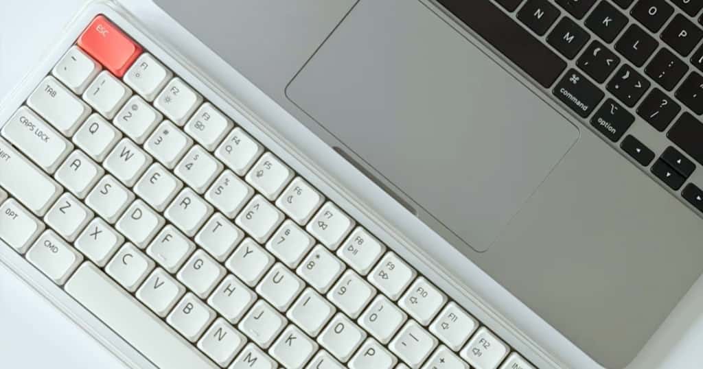 Se muestra un teclado inalámbrico blanco colocado junto a un MacBook sobre una mesa para ilustrar por qué se utiliza un teclado externo inalámbrico en un MacBook